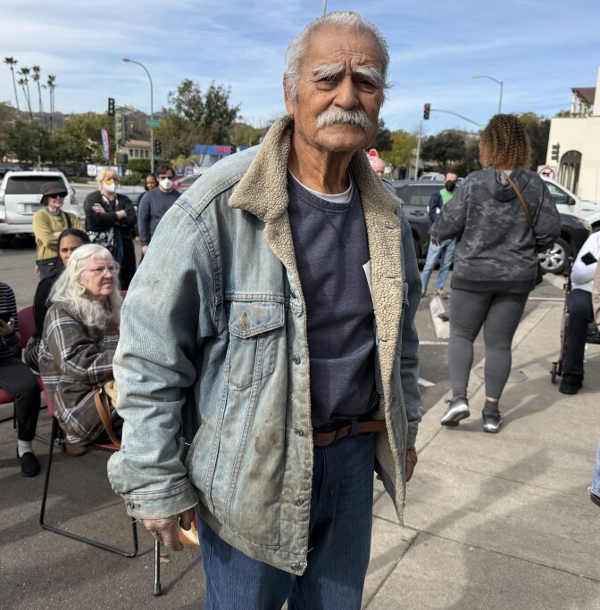 A man with light brown skin, short white hair and a white mustache stands outside a building. He wears a light blue denim jacket that has dirt or soil on the front and at the collar. He's also wearing blue jeans and a navy blue sweatshirt.
