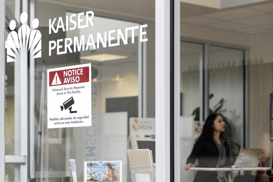 The exterior of a building with glass windows. The building says "Kaiser Permanente" in white lettering in the top right of the image. A woman is pictured in the background.