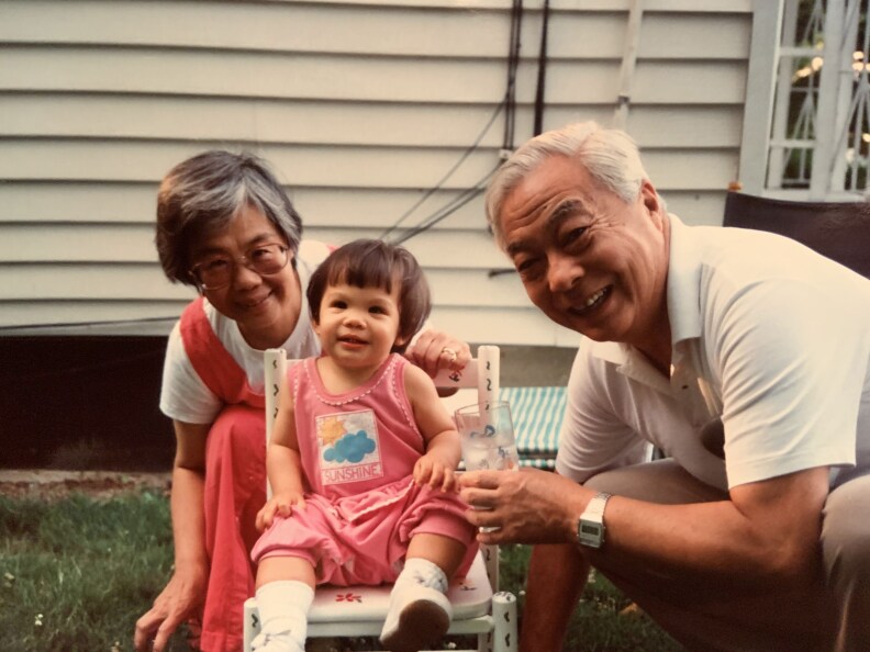 An Asian woman kneels behind a young baby who is sitting in a white kid's chair. Adjacent to them is an older Asian man holding a glass and wearing a watch. They are all in front of a white colored building.