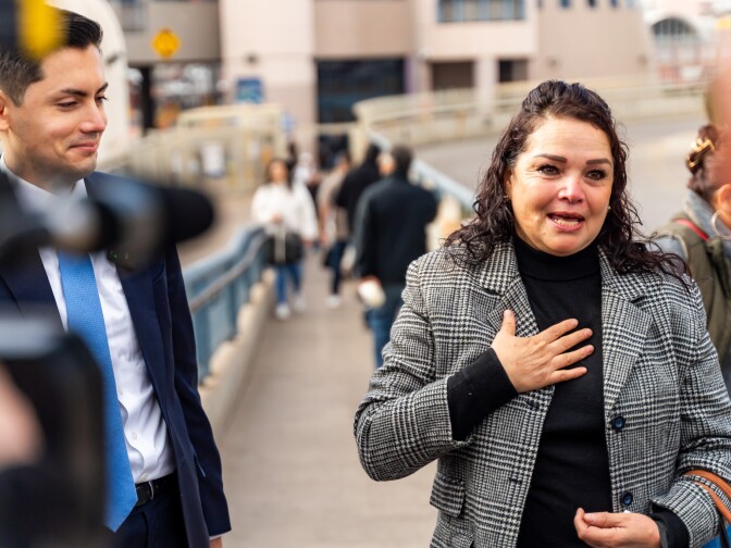 A young man in a suit stands next to a middle-aged woman in a blazer, who speaks to reporters. 