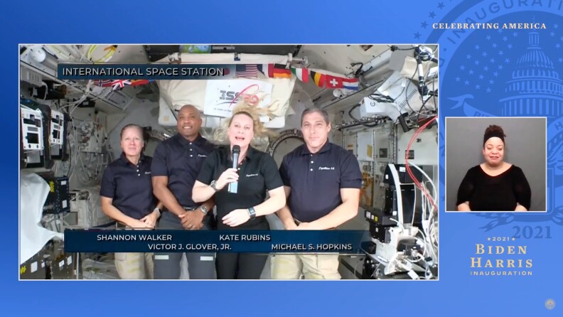 A screengrab from a live video stream depicts four astronauts (two men and two women) aboard the International Space Station. A female astronaut holds a microphone and appears to be talking, while an American Sign Language interpreter signs on the right side of the screengrab.