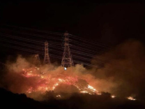 A burning hillside with powerlines within the flames is seen with smoke rising into the night sky.