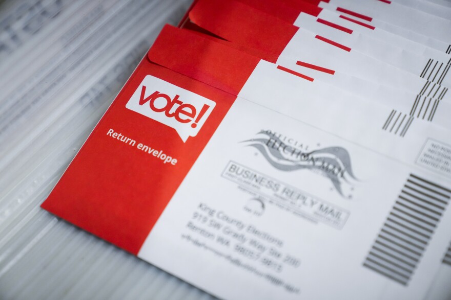 White envelopes with the left third of the envelopes colored red. These mail in ballots have the words "vote" and "return envelope" printed on the red portion of the envelope. There's also a blurred address on the white portion