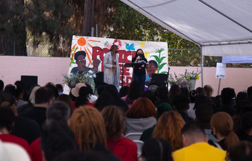 Two speakers in the distance stand on a stage in front of a hand-painted banner that says "Police Free LAUSD." The speakers are addressing a large group of students seated under a white tent.