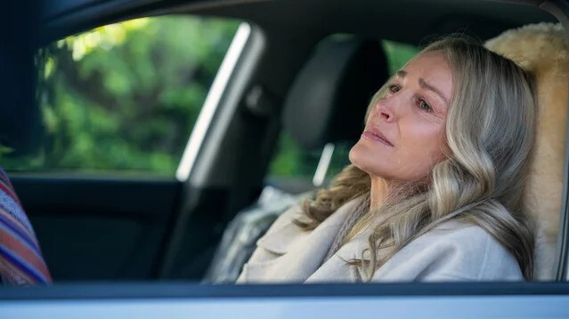 A woman who appears to be white and in her 50s or 60s slouches in the driver's seat of a car, looking anguished. She has shoulder-length blond hand and wears a camel colored coat.
