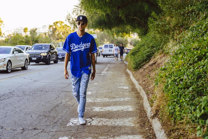 A person wearing a Dodger Blue jersey trudges up the pedestrian path to the stadium, moving much faster than the long line of vehicles nearby in bumper-to-bumper traffic, also waiting to get to the ballpark.