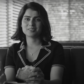 Anaita Wali Zada sits in a booth at a diner. She holds a half eaten sandwich in her hand, a glass of something to her right. She smiles softly at whoever is sitting in the opposite booth to her.