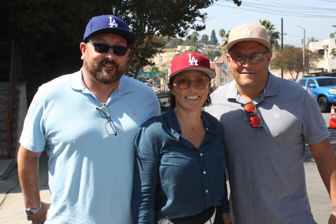 A group of three people stand, two men and a woman, stand facing the camera, wearing sunglasses and Dodgers hats in different colors