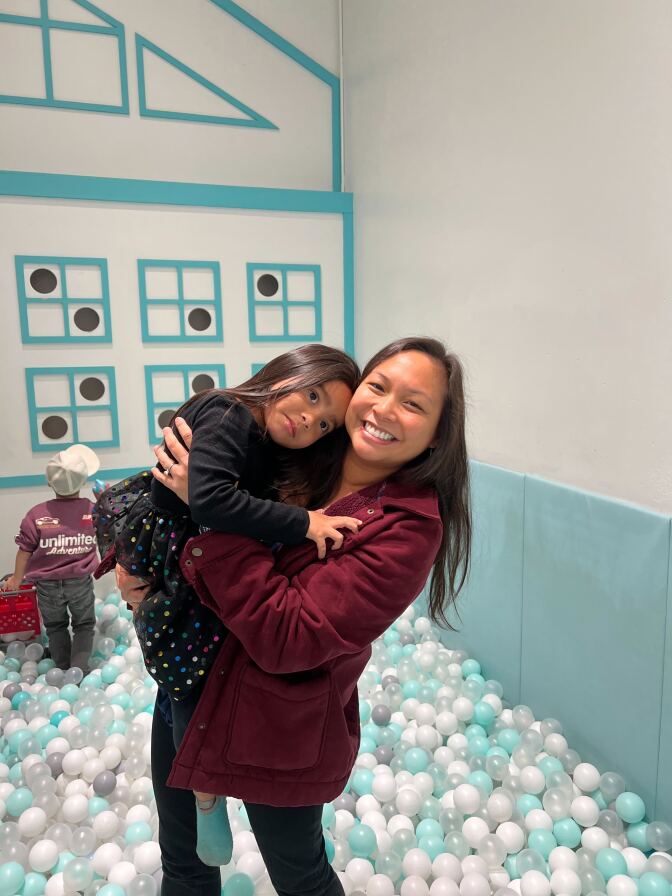 A female with olive skin tone and dark hair wearing a maroon hoodie carries a child wearing a black dress. 