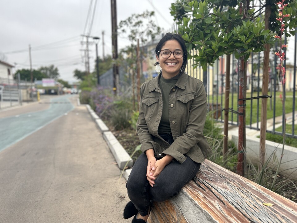 A woman with black hair and light brown skin wearing black glasses, a green shirt and jacket and black pants smiles for the camera. She sits  on a wood bench with trees and a well-paved alley in the background. 