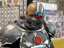 A brown-skinned man in a Cyborg cosplay outfit, with a robotic covering over his mouth (and a COVID-19 mask peeking through). He stands in front of a brightly colored convention booth in soft focus in the background.