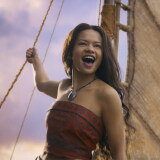 A female-presenting person wearing a terracotta strapless top and a necklace stands holding the rope of a sail. The sail and a cloudy sky are visible in the background.
