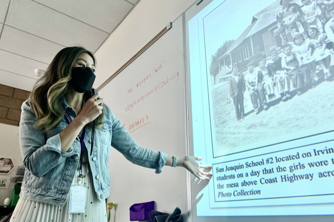 An Asian American woman with ombre dyed hair and wearing a jean jacket stands before a white board onto which a black-and-white photo of Japanese American schoolchildren has been projected. 