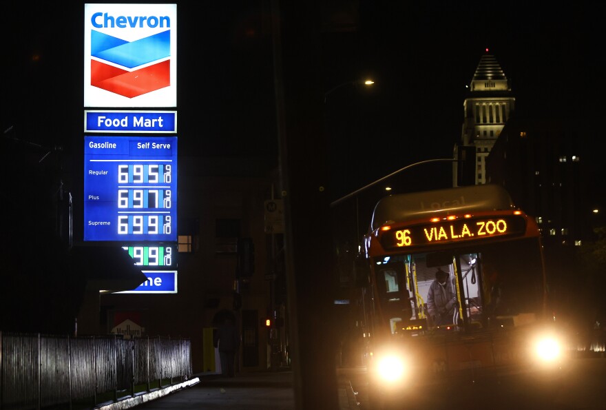A bus with the lighted sign 96 VIA L.A. ZOO passes by a Chevron station where the price of a gallon of regular gas is $6.95. L.A. City Hall is lighted in the background.