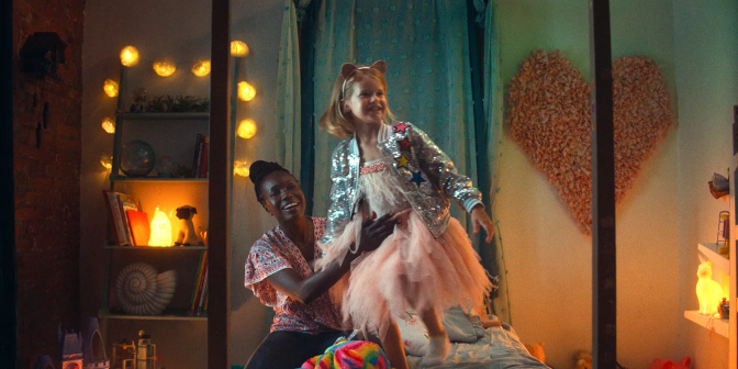 Anna Diop and Rose Decker are in a young girl's bedroom. Diop sits on the small single bed as Decker stands on top of it, Diop gentling holding her by the waist to help her balance. Both are smiling. Decker wears a pink tulle dress with a silver sequenced jacket. Diop wears a blue jeans and a red patterned top with short, flowy sleeves. The bedroom they are in is illuminated by the various string lights and lamps, giving the room a warm yellow glow. The shelves in the room are filled with books and knick-knacks. A blue curtain hangs from the wall behind Diop and Decker and a giant floral heart hangs on the wall beside it. 