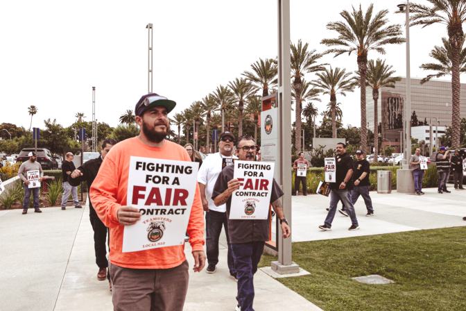 Men walk in front of county buildings holding signs that say, "Fighting for a fair contract." 