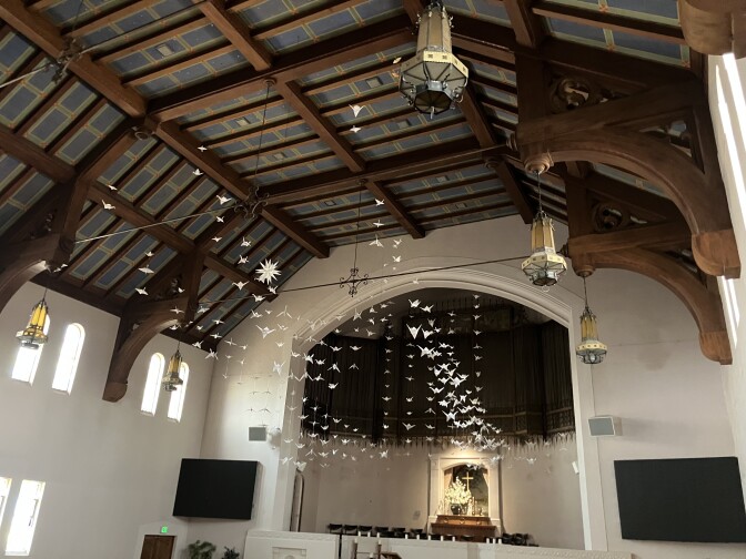 A large room with high ceilings and white walls. Lights hang from the ceiling, which has wooden boards. A cross and altar can be seen at the front of the room. Dozens of paper cranes hang from the ceiling, creating the illusion that they are floating.