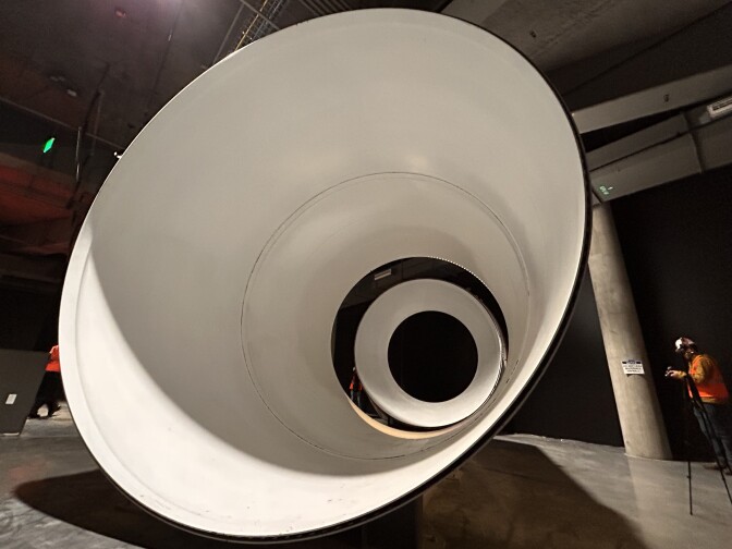 A massive stark-white circular metal object is displayed while resting on the ground. The object is thin and open in the middle, allowing people to pass through.