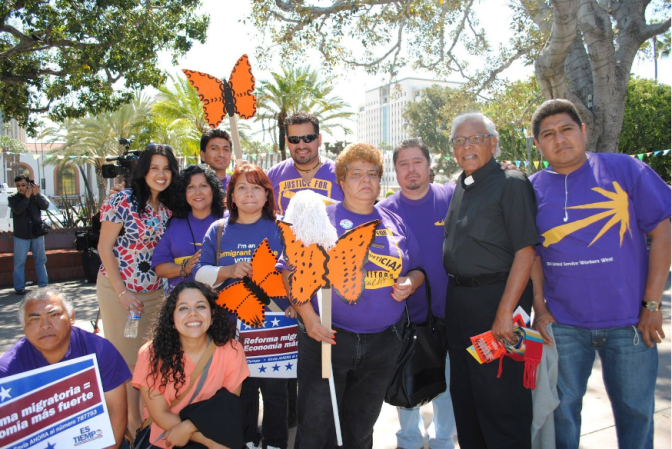 Close to a dozen people of varying shades of medium skin tone stand in a group, with some wearing purple shirts with yellow graphics. One man is wearing what looks like a black clergy shirt. Several carry what looks like cardboard cutouts of Monarch butterflies, mop heads, and signs with slogans in spanish. They stand in what looks like a plaza outside.