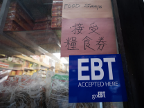 A sign that reads "EBT Accepted Here" hangs in the window of a shop. It hangs below two pink signs, one with Chinese characters and aonther with the words "Food Stamps." Plastic wrapped goods are lined up against the window.