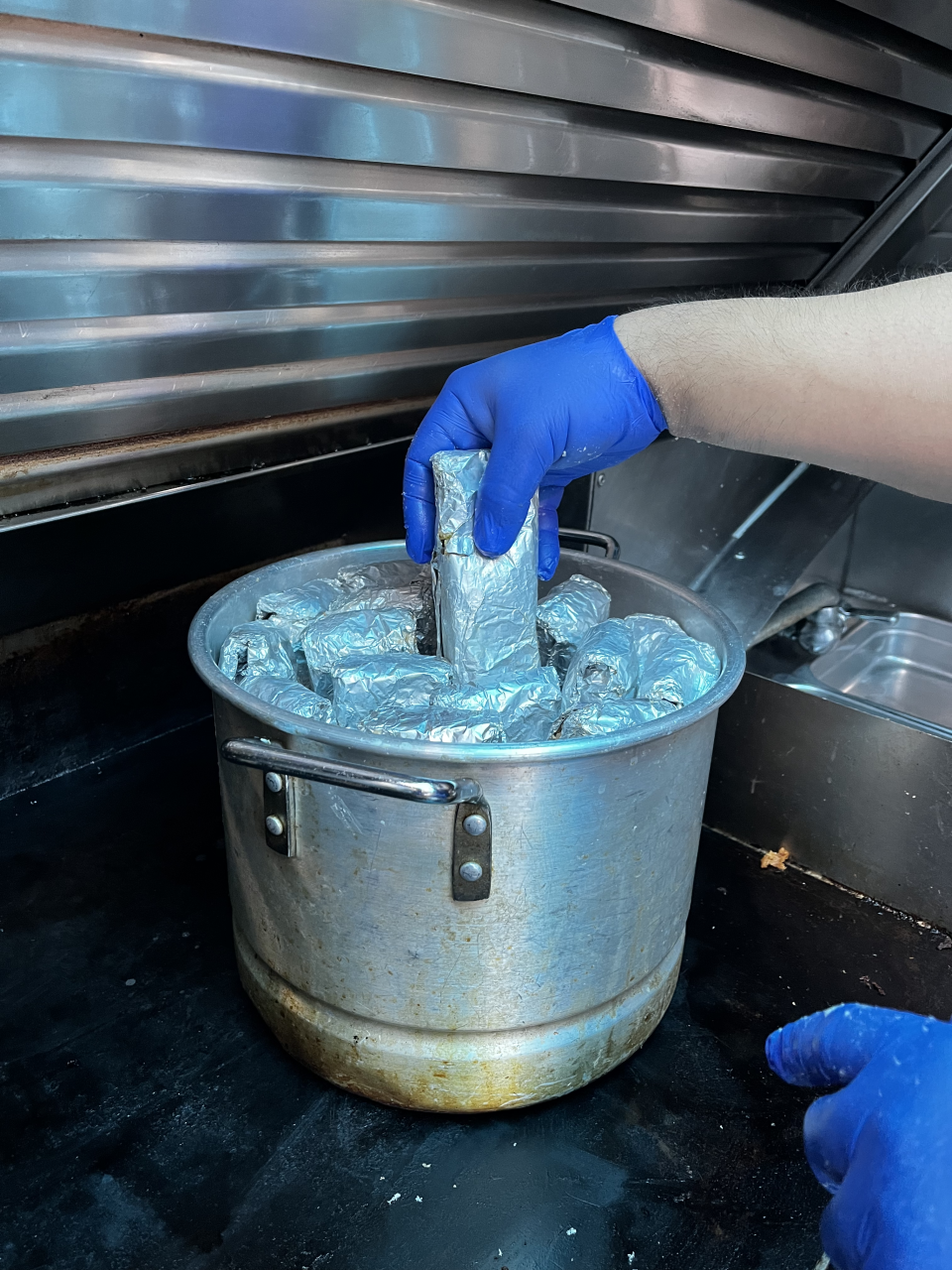 A gloved hand places a tamal wrapped in foil into a metal pot inside of the food truck