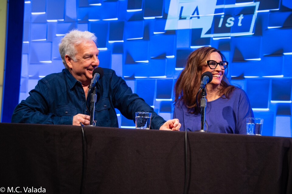 Marc Summers, a white man and Michaela Watkins, a white woman with glasses, smiling behind microphones at the panelist table looking to the right with the LAist logo on the wall behind
