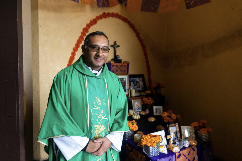 A priest stands in front of a traditional Dia e los Muertos altar.
