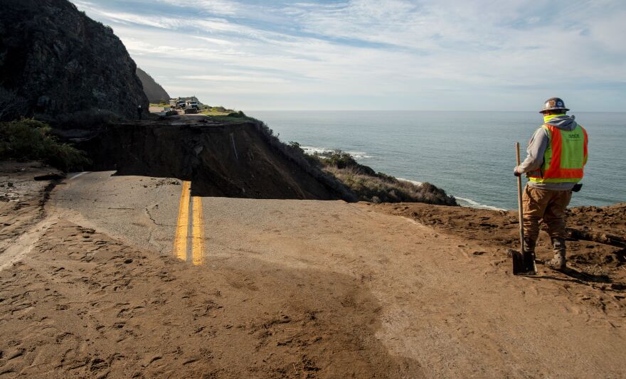 A person wearing a safety vest and carrying a shovel stands near a gaping hole in Highway 1 where the road collapsed into the ocean.