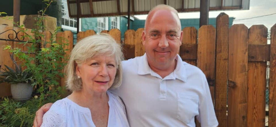 A white woman and white man stand with their arms around each other in front of a wood fence.