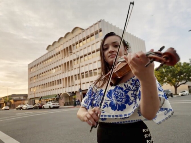 A female presenting person wears a blue, floral top. She plays the violin. She is standing in front of a white office building with dozens of windows.