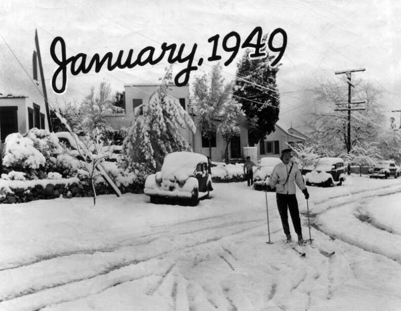 A black and white image of a person skiing down a snow covered street. There are cars parked around covered in snow and trees filled with ice. January, 1949 is on top of the image.