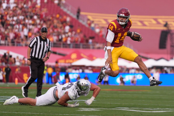 A football player in a cardinal and gold "SC" uniform, number 14, leaps through the air holding the ball while a defender in a white uniform and silver helmet dives at the turf below him.