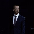 A male-presenting person with a strawberry blonde buzz-cut stands in a dark room wearing a white button-up dress shirt, a gray tie, and a navy blue suit jacket. There is a yellow curtain behind him on his right, and the upper part of a leather chair is visible in the bottom right corner of the image.