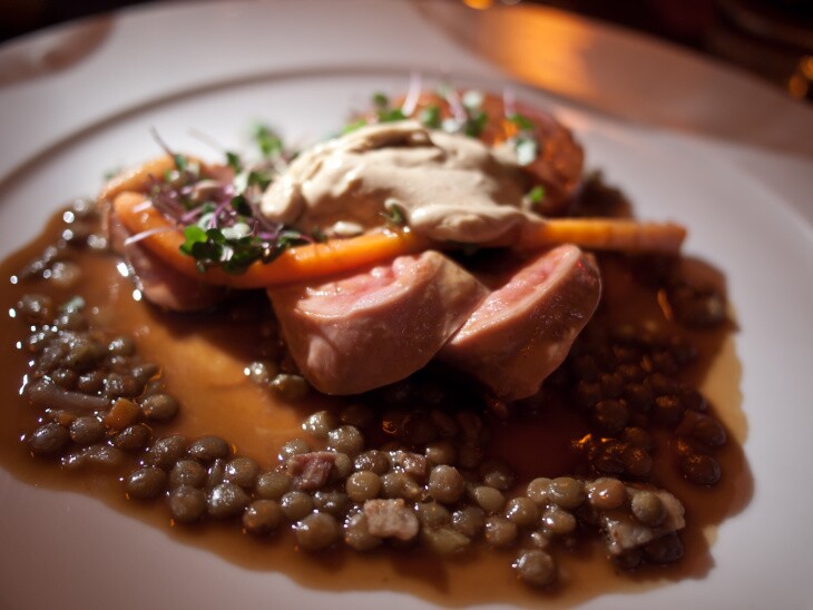 A close up photo of a white dinner plate holding a soupy lentils topped off with sliced sausage and slivers of carrots, along with other veggies.
