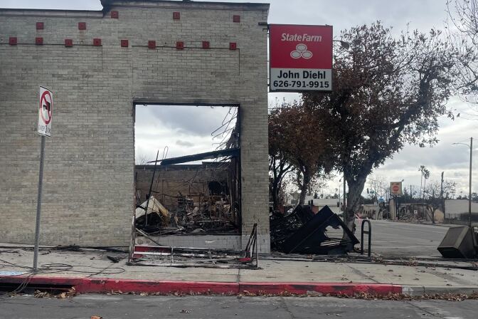 Ruins of a burned building with a State Farm sign outside. The off-white brick exterior of the building remains standing. The sign outside reads "State Farm John Diehl 626-791-9915." Wreckage of other buildings is visible in the background against gray skies.