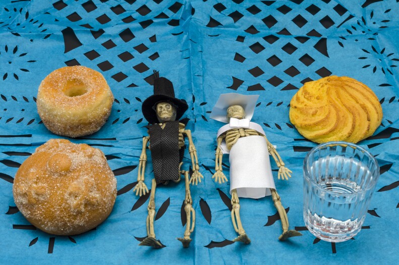 Plastic skeletons dressed as bride and groom in an offering altar for mexican day of the dead with blue cut paper, bread and mezcal