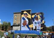 A mural featuring a baseball player on two different panels of a mural. A crowd is below looking at it.