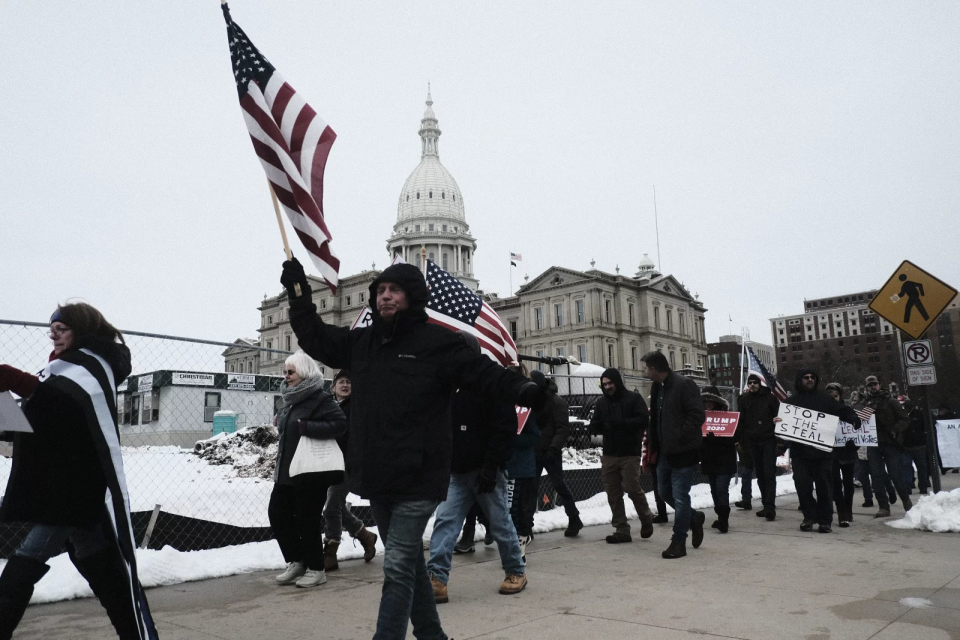 People in front of state official buildings hold signs and wave American flags