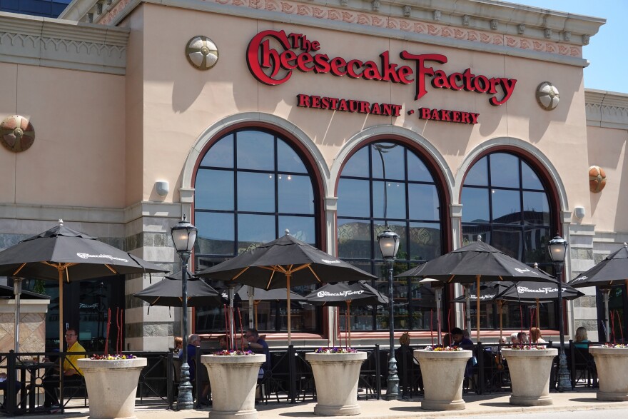 A beige multistory building with red signage that spells out "Cheesecake Factory" is pictured in the daylight surrounded by tables and chairs.