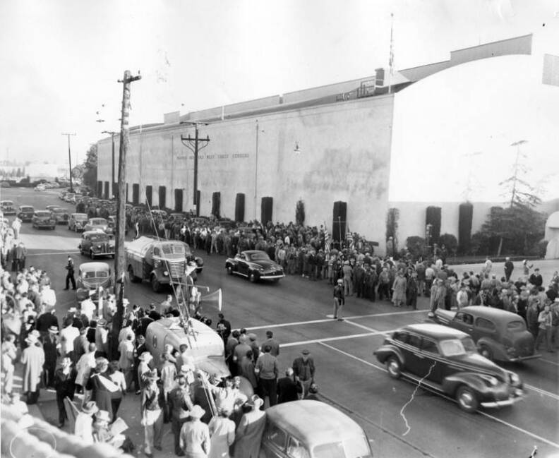 The picketers are walking the entire length of the building. Policemen can be seen on both sides of the street. Cars are driving down the street and one car with loudspeakers on top is parked on the opposite side. An American flag flies over the building. The wording on the building reads "Warner Brothers West Coast Studios."
