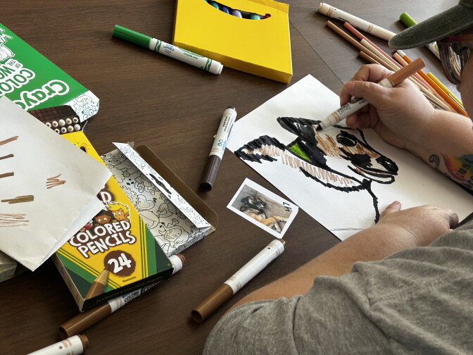 A hand colors a marker drawing of a dog, referencing a small photo, surrounded by markers, crayons and colored pencils on a table.