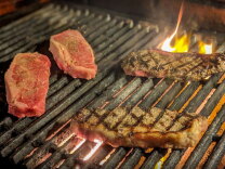 Four raw and grilled steaks cooking on a flaming charcoal grill, two seared with grill marks and two still raw.