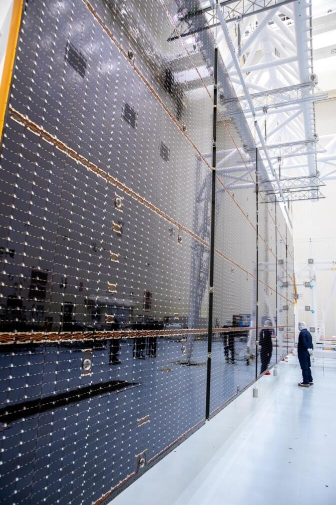 A man wearing a black protective full-body suit and white gloves and mask is standing in a stark white laboratory. Several enormous black solar panels are positioned in front of him, towering over the space.