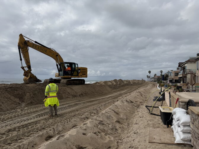 A man in reflective clothing watches a backhoe move sand on a beach under cloudy skies.