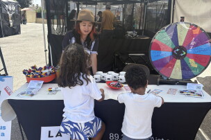 An outdoor photograph captures a community booth hosted by LAist, set up under a white canopy tent on an asphalt surface. A staff member wearing a brown wide-brimmed hat stands behind a table draped with a black tablecloth that features the LAist logo and text reading "89.3 LAist.com". Two young children with curly dark hair, viewed from behind, stand at the front of the table looking at the booth's activities. On the table sit white mugs, a red bowl filled with markers, informational flyers, and an orange basket holding pens. To the right, a large colorful prize wheel is positioned for a game, displaying sections labeled with items like "sippy cup" and "pencil sharpeners".