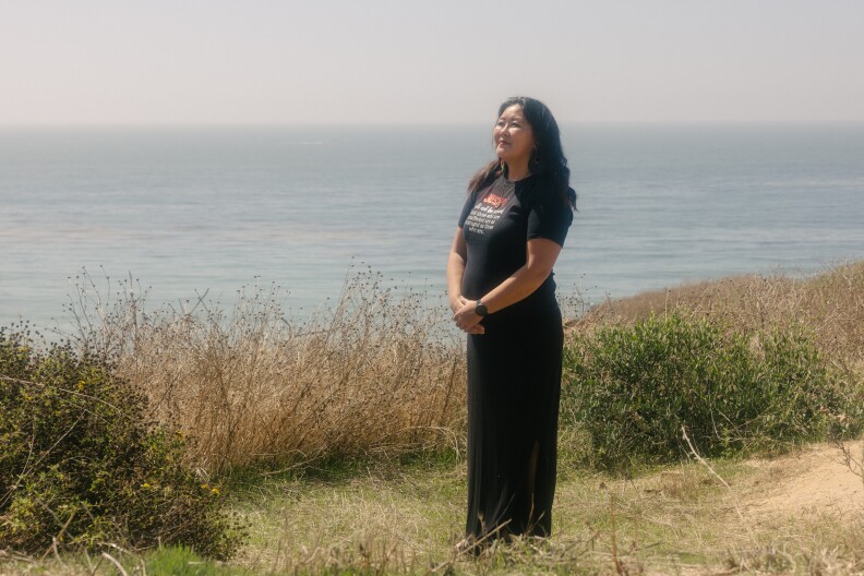 An Asian woman with long dark hair wearing a black t-shirt and pants and flip flops stands near an ocean side cliff looking out at the sun.