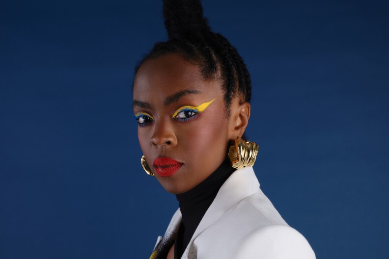 Lauryn Hill looks at the camera wearing a black turtleneck, a white suit coat, gold earrings and yellow and blue eye shadow.