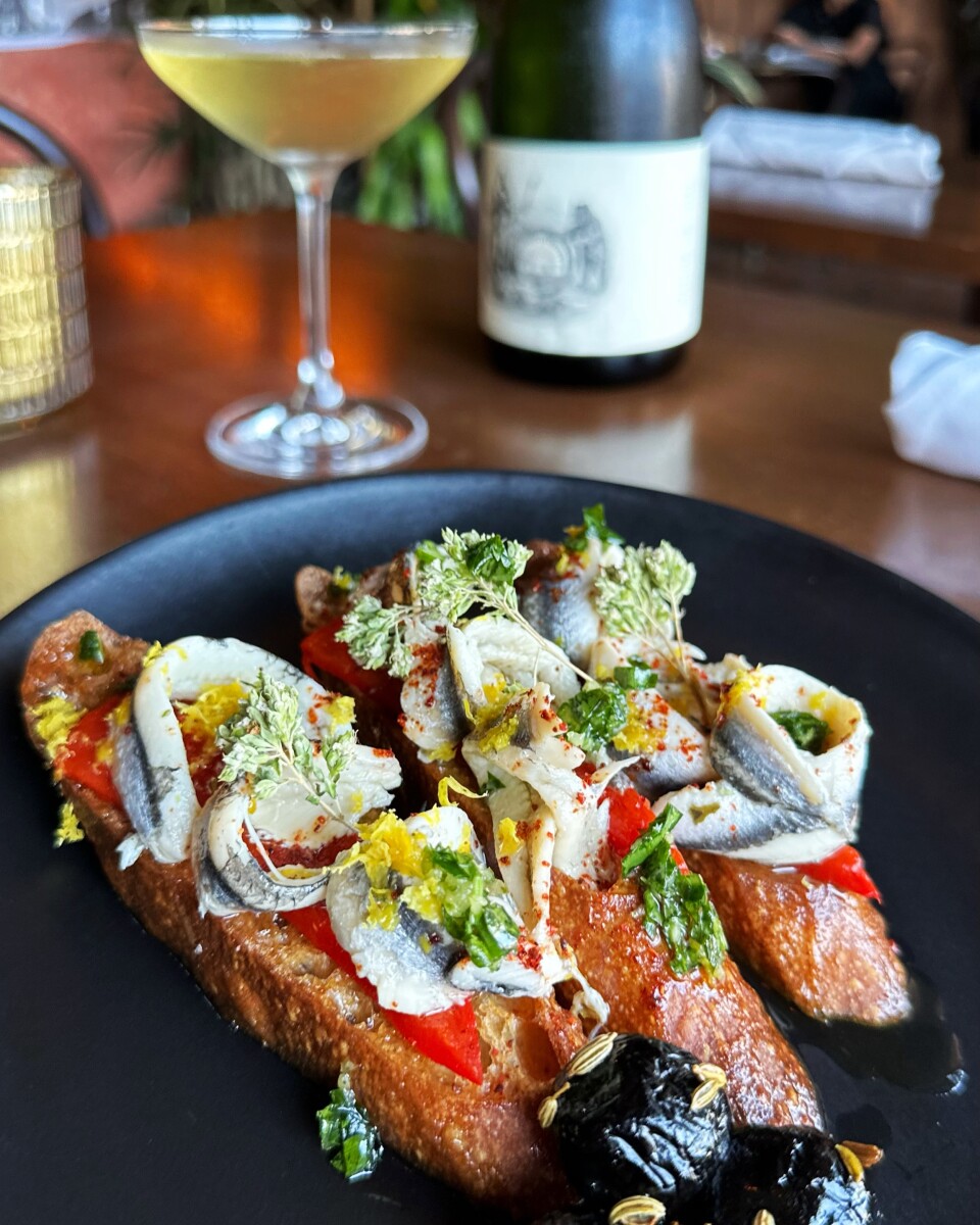 A plate with three bias-cut bread slices topped with red pepper, fresh white anchovy, herbs, and spices. A glass of white wine and a wine bottle are in the background.