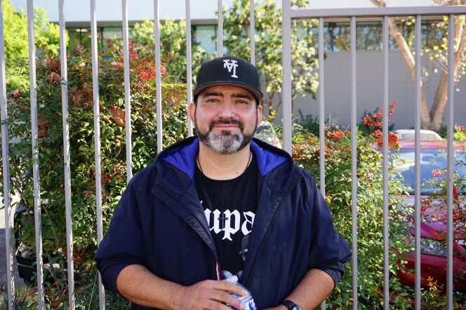 A man with medium-light skin tone stands in front of a fence with a plant poking through. He wears a black baseball cap with an upside down "LA." He has a gray beard. He smiles lightly at the camera.