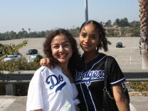 Two women pose for the camera. The woman on the right is wearing a white Dodger jersey and has brown curly hair. The woman on the left is wearing a black Dodgers jersey with black curly hair. 